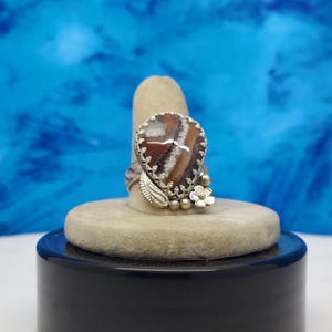 May include: A silver ring with a heart-shaped stone set in the center. The stone is a mix of brown, white, and gray colors. The ring is decorated with silver flowers and leaves.