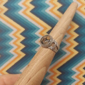 May include: A silver ring with a large, round, patterned stone. The stone has a mix of tan, brown, and black colors. The ring has a decorative, openwork design around the stone and on the band.