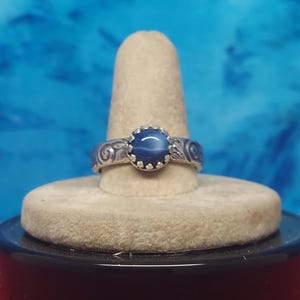 May include: A silver ring with a large, oval, dark blue gemstone at the center. The ring has a decorative, swirling pattern on the band and a scalloped setting around the gemstone. The ring is displayed on a beige ring holder.