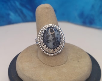 May include: A silver ring with a large, round, gray and white stone. The stone is set in a decorative silver bezel with a beaded edge and a crown-like design. The ring is displayed on a beige ring holder.