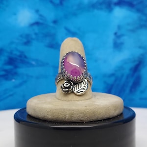 May include: A silver ring with a purple and pink agate stone set in a floral design. The ring is on a display stand.