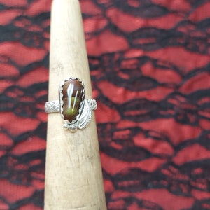 May include: A silver ring with a brown and green oval gemstone set in a silver band. The band is decorated with a silver leaf design.
