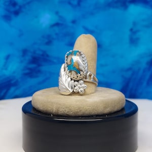 May include: A silver ring with a turquoise stone set in a floral design. The ring is on a display stand.