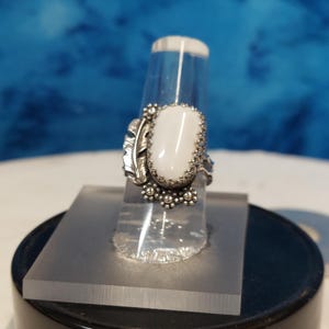 May include: A silver ring with a white oval stone and a leaf design. The ring is on a clear display stand.