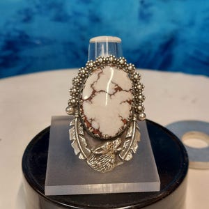 May include: Oval-shaped ring with a white and brown stone set in a silver-colored metal. The ring features a detailed design with feathers and a wolf's head. The stone is surrounded by small, round embellishments.