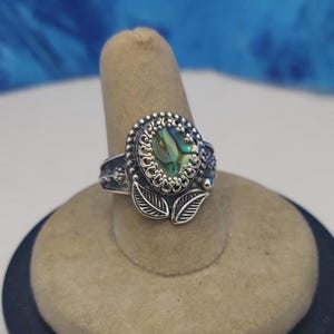 May include: A silver ring with an oval abalone shell center. The ring features a detailed design with a beaded border, leaf accents, and ornate side detailing. The abalone shell displays iridescent green and blue hues.