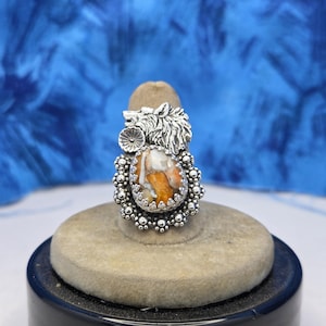 May include: A silver ring featuring a wolf design and a large, oval-shaped stone with orange and white patterns. The stone is surrounded by a beaded silver setting. The ring is displayed on a beige ring holder.