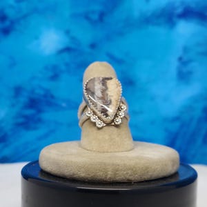 May include: A silver ring with a teardrop-shaped stone set in the center. The stone is a light brown color with white and gray swirls. The ring is on a gray ring display stand.