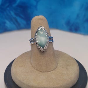 May include: A silver ring featuring a light blue oval gemstone. The gemstone is set in a detailed filigree setting. The ring is displayed on a neutral-toned ring holder against a blue background. The ring's band has a decorative design.