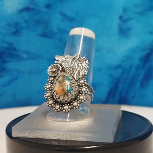 May include: A silver ring with a wolf head design and a turquoise and orange gemstone. The ring is set on a white display stand.