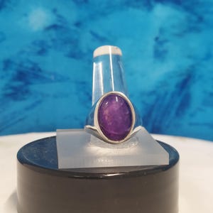 May include: A silver ring featuring a large, oval-shaped, deep purple gemstone. The ring is set in a simple, polished silver band. The gemstone is the focal point, showcasing its rich color and smooth surface. The ring is displayed on a clear stand against a blue background.
