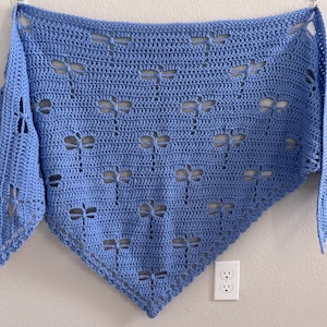 May include: A triangular crochet shawl in a light blue color featuring a repeating openwork dragonfly pattern throughout the fabric. The shawl has a pointed bottom edge and a straight top border, showcasing intricate yarn work and stitch detail.