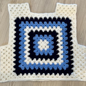 May include: A handmade crochet vest in a granny square pattern. The vest features a square design with concentric squares of white, light blue, dark blue, and white. The vest has shoulder straps and a square neckline.