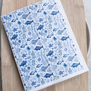 May include: A white rectangular tea towel with a blue fish and aquatic plant pattern. The design features various fish and plant illustrations in shades of blue, creating a cohesive and decorative look. The tea towel is placed on a wooden chopping board.