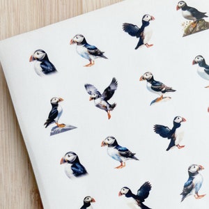 May include: A sheet of stickers featuring colorful illustrations of puffins in various poses. The puffins are depicted with black and white markings, orange beaks, and white faces. The stickers are printed on white paper.