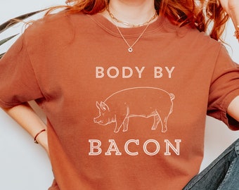 Könnte beinhalten: Eine Frau trägt ein rostfarbenes T-Shirt mit dem Text "BODY BY BACON" und einer weißen Umrandung eines Schweins. Sie trägt eine blaue Jeans und weiße Turnschuhe.