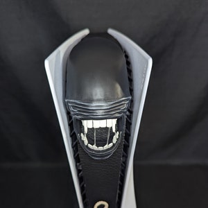 May include: A white and black gaming console with a black alien head sculpture on top. The alien head has a large open mouth with sharp teeth.