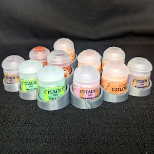 May include: A collection of Citadel Dry paint pots in various colors, including orange, pink, and green, with clear lids. The pots are arranged in gray holders, creating a display of miniature painting supplies.