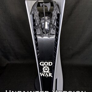 May include: A white and black PlayStation 5 console with a decorative panel featuring the "God of War" logo and weapon designs. The text "UNPAINTED VERSION" is displayed at the bottom.