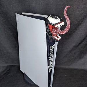 May include: A white PlayStation 5 console with a black Venom figure attached to the side. The figure has a red tongue and is in a menacing pose.