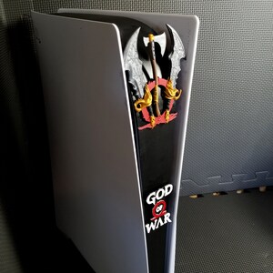 May include: A white and black PlayStation 5 console with a decorative panel featuring the "God of War" logo and a detailed axe design. The panel is red, gold, and gray.