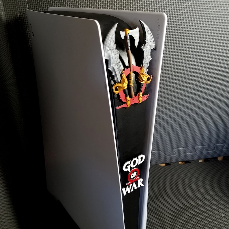 May include: A white and black PlayStation 5 console with a decorative panel featuring the "God of War" logo and a detailed axe design. The panel is red, gold, and gray.