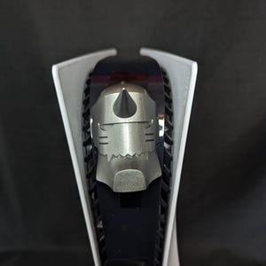 May include: A silver-colored decorative piece, shaped like a helmet, is placed on a black and white game console. The helmet has a pointed top and detailed facial features. The console has a sleek, modern design.