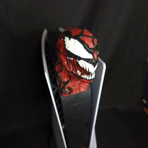 May include: A red and black Venom symbiote head figurine with white eyes and sharp teeth. The figurine is mounted on a black leather base with a white and red spider logo.