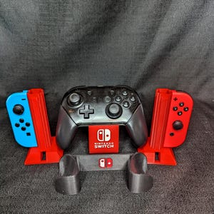 May include: A black Nintendo Switch Pro controller and red and blue Joy-Con controllers are displayed in a 3D-printed stand. The stand is red and gray, with the Nintendo Switch logo visible.