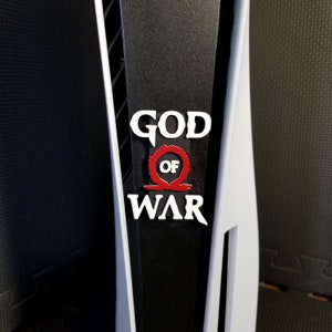 May include: A close-up of a black and white PlayStation 5 console with the "God of War" logo. The logo features white text with a red symbol in the center. The console has a sleek, modern design.