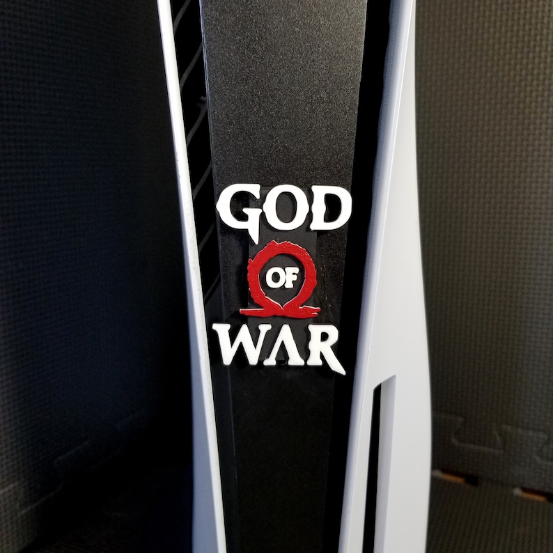 May include: A close-up of a black and white PlayStation 5 console with the "God of War" logo. The logo features white text with a red symbol in the center. The console has a sleek, modern design.