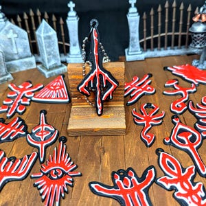 May include: A collection of black and red pendants and charms with various designs, including a key chain. The pendants are arranged on a wooden surface with miniature tombstones and a decorative lantern in the background. The designs have a gothic theme.