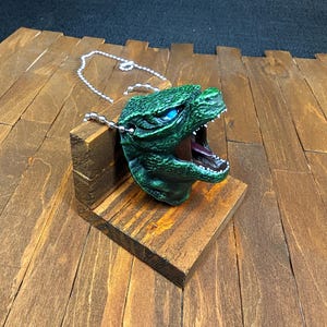 May include: A green and blue monster head necklace with a silver chain. The monster has a large mouth and sharp teeth. The necklace is displayed on a wooden stand.