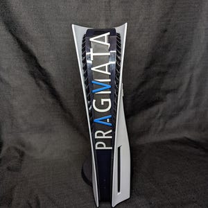 May include: A white and black gaming console with the word "PRAGMATA" in white and blue lettering. The console is standing upright on a black base against a dark gray background.