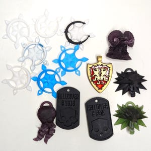 May include: An assortment of fantasy-themed pendants and charms in various colors. Includes clear, blue, and black starburst shapes, a black ram, a shield, a black spiky charm, a green charm, and black dog tags with a skull and the text "HELLDIVER #5918".
