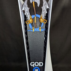 May include: A white and black PlayStation 5 console with a custom faceplate featuring the God of War logo and imagery. The faceplate includes a blue and gold design with the game's iconic weapons.