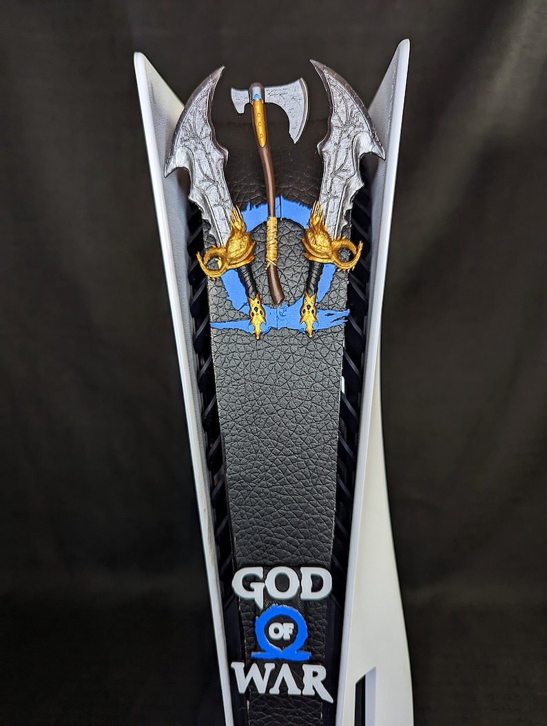 May include: A white and black PlayStation 5 console with a custom faceplate featuring the God of War logo and imagery. The faceplate includes a blue and gold design with the game's iconic weapons.