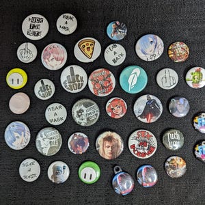 May include: A collection of various round pinback buttons with diverse designs and text. Some buttons feature anime-style characters, pizza slices, and the text "FUCK THE ELITE", "WEAR A MASK", and a middle finger graphic. The buttons are in various colors.