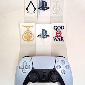 May include: A white and black PlayStation 5 controller is displayed below six game logo designs. The logos are on white paper strips. The logos include the PlayStation logo, Assassin's Creed, and God of War.