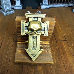 May include: A gold-colored metal pendant in the shape of a cross with a skull in the center. The pendant is on a chain and is sitting on a wooden surface.
