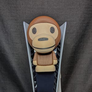 May include: A wooden monkey-faced figure with a brown head, tan face, and brown body. The figure is designed to fit on a white and black game console. The monkey has large, round eyes and a smiling mouth.
