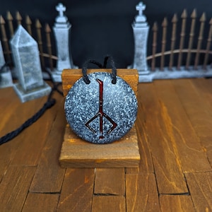 May include: A round, speckled pendant with a black cord. The pendant has a dark gray base with white speckles and a red symbol in the center. The pendant is displayed on a small wooden stand.