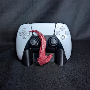 May include: A white and black PlayStation 5 controller with a red, textured, tongue-like object attached. The controller rests on a black stand against a dark background. The controller has buttons and a directional pad.