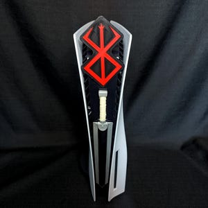 May include: A white gaming console with a red and black emblem on top. A silver sword with a tan handle is in the center. The console has a sleek, modern design, with a black background.