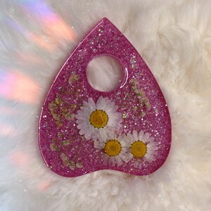 May include: Pink glitter resin planchette with a hole in the center and three white pressed flowers.