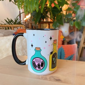 May include: White ceramic mug with a black handle. The mug has a design of two potion bottles with labels that read "Ghost Tears" and "Batwing". The bottles are surrounded by pink and black stars.