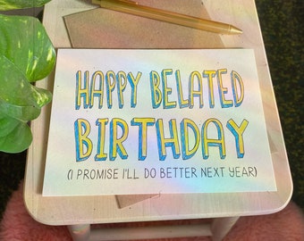 May include: A holographic card with the text "Happy Belated Birthday (I promise I'll do better next year)" in yellow and blue lettering.