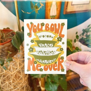 May include: A white card with the text "You bowl me over" in orange and green letters. The card features a stack of three green and white bowls with floral patterns.