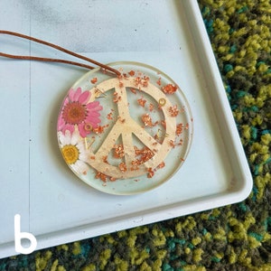 May include: A round, clear resin ornament with a peace sign design, copper glitter, and pressed flowers. The ornament is attached to a brown leather cord.