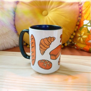 May include: White ceramic mug with a dark blue interior and handle. The mug features illustrations of various baked goods in orange, including a baguette, croissant, and loaf of bread. The mug is sitting on a wooden surface.